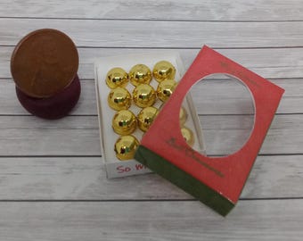 May include: A collection of small, gold-colored Christmas ornaments in a white box, a red card with a circular cutout, and a brown penny on a dark purple base, all on a light wood-grain surface.