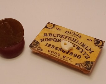 May include: A miniature Ouija board with a planchette and a brown, circular object. The board is tan with black lettering and a crescent moon design. The planchette is off-white.