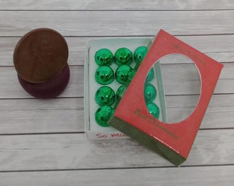 May include: A brown and purple decorative item next to a box of green glass ornaments. The box has a red cover with a circular cutout. The ornaments are small, round, and shiny. The background is a light wood grain.