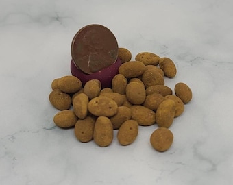 May include: A pile of small, oval-shaped, golden-brown snacks. A U.S. penny rests on a small, dark purple object, with the snacks scattered around it. The snacks have a textured surface and are similar in size and shape.