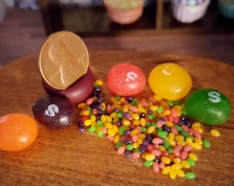 May include: A collection of colorful candies, including orange, red, yellow, and green, scattered on a wooden surface. A penny is propped up on a small, dark purple candy. A variety of smaller, jelly bean-like candies are also present.