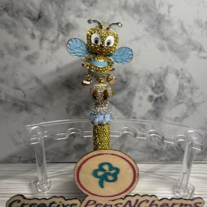 May include: A decorative pen featuring a jeweled bee design with yellow, blue, and gold accents. The pen has a gold glitter barrel and a wooden base with a clover design. The base also has the text "Creative Pens N Charms" and "Pens that Spark Joy".