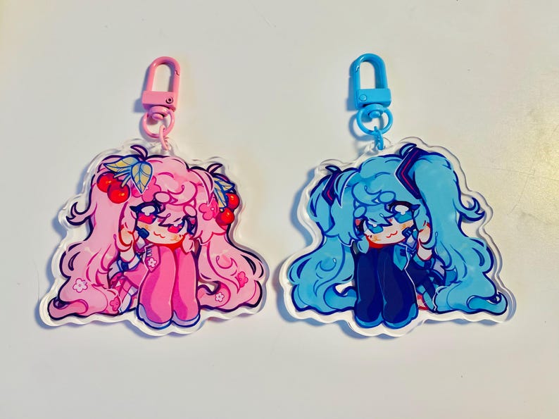 Pu&ograve; includere: Due ciondoli in acrilico con illustrazioni di personaggi in stile anime. Uno &egrave; rosa con ciliegie, l'altro blu. Ogni ciondolo ha una chiusura colorata. Adatto per portachiavi.