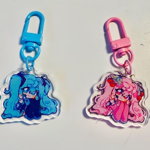Pu&ograve; includere: Due portachiavi in acrilico con personaggi in stile anime. Un portachiavi ha un personaggio blu con capelli blu e un fermaglio blu. L'altro portachiavi ha un personaggio rosa con capelli rosa e un fermaglio rosa. Entrambi i portachiavi hanno un bordo trasparente.