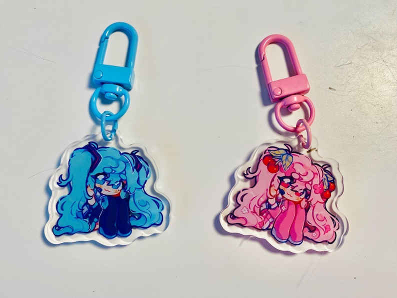 Pu&ograve; includere: Due portachiavi in acrilico con personaggi in stile anime. Un portachiavi ha un personaggio blu con capelli blu e un fermaglio blu. L'altro portachiavi ha un personaggio rosa con capelli rosa e un fermaglio rosa. Entrambi i portachiavi hanno un bordo trasparente.
