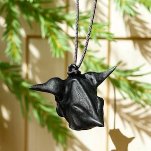 May include: A black pendant necklace featuring a textured, stylized design. The pendant is suspended from a dark cord. The design has a unique shape with pointed elements. The background includes green foliage and soft lighting.
