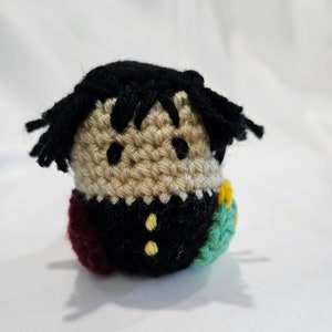 May include: A handmade crochet doll with black hair and a tan face. The doll is wearing a black outfit with yellow accents, and a green and burgundy section on the side. The doll is approximately 3 inches tall.