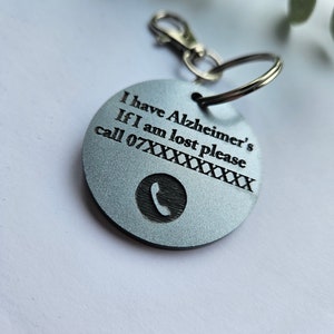 Puede incluir: Un llavero plateado con una etiqueta redonda que dice "Tengo Alzheimer Si me pierdo, por favor llama al 07XXXXXXXXX" con un icono de teléfono.