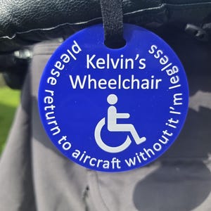 May include: A circular blue plastic tag with a black strap featuring the white text Kelvin's Wheelchair and please return to aircraft without it I'm legless, along with a white wheelchair accessibility symbol centred on the surface.
