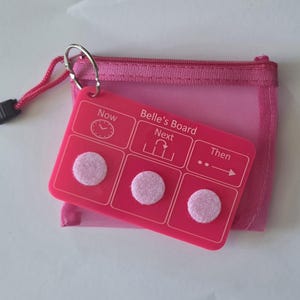 May include: A bright pink rectangular plastic board labelled Belle's Board with three sections titled Now, Next, and Then, each featuring a light pink circular velcro dot. The board is attached by a metal ring to a matching pink mesh zip-up pouch.