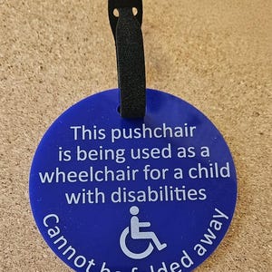May include: A blue circular tag with white text and a wheelchair symbol. The text reads: "This pushchair is being used as a wheelchair for a child with disabilities. Cannot be folded away." A black loop is attached.