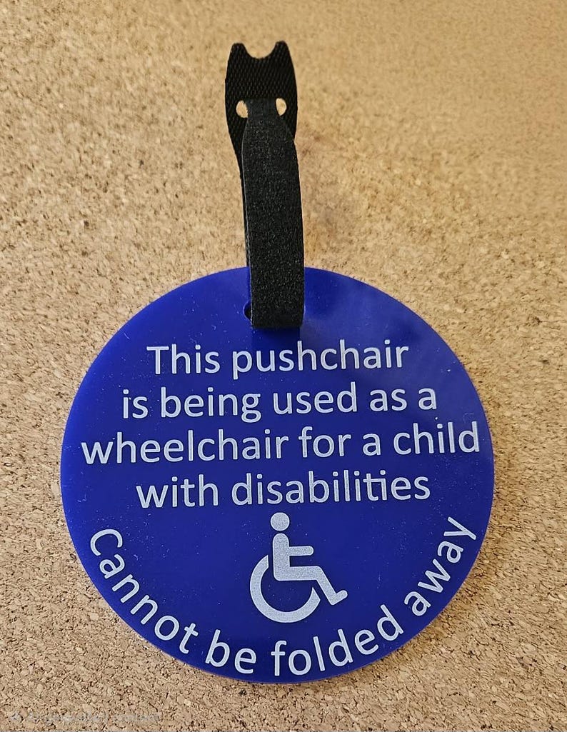 May include: A blue circular tag with white text and a wheelchair symbol. The text reads: "This pushchair is being used as a wheelchair for a child with disabilities. Cannot be folded away." A black loop is attached.