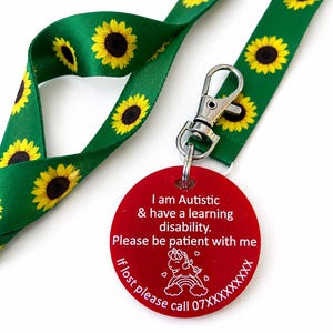 Peut inclure: Lanière verte ornée de tournesols jaunes et d'une étiquette ronde rouge. L'étiquette indique : "Je suis autiste et j'ai des difficultés d'apprentissage. Soyez patient avec moi. En cas de perte, appelez le 07XXXXXXX". Avec un fermoir argenté.