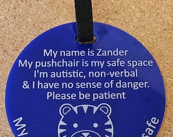 May include: A circular blue plastic tag with a black strap. The text reads: My name is Zander, My pushchair is my safe space, I'm autistic, non-verbal & I have no sense of danger. Please be patient. Below is a tiger graphic and My pushchair keeps me safe.