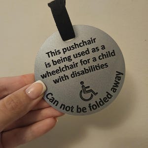 May include: A silver, circular tag with black text and a wheelchair symbol. The text reads: "This pushchair is being used as a wheelchair for a child with disabilities. Can not be folded away."