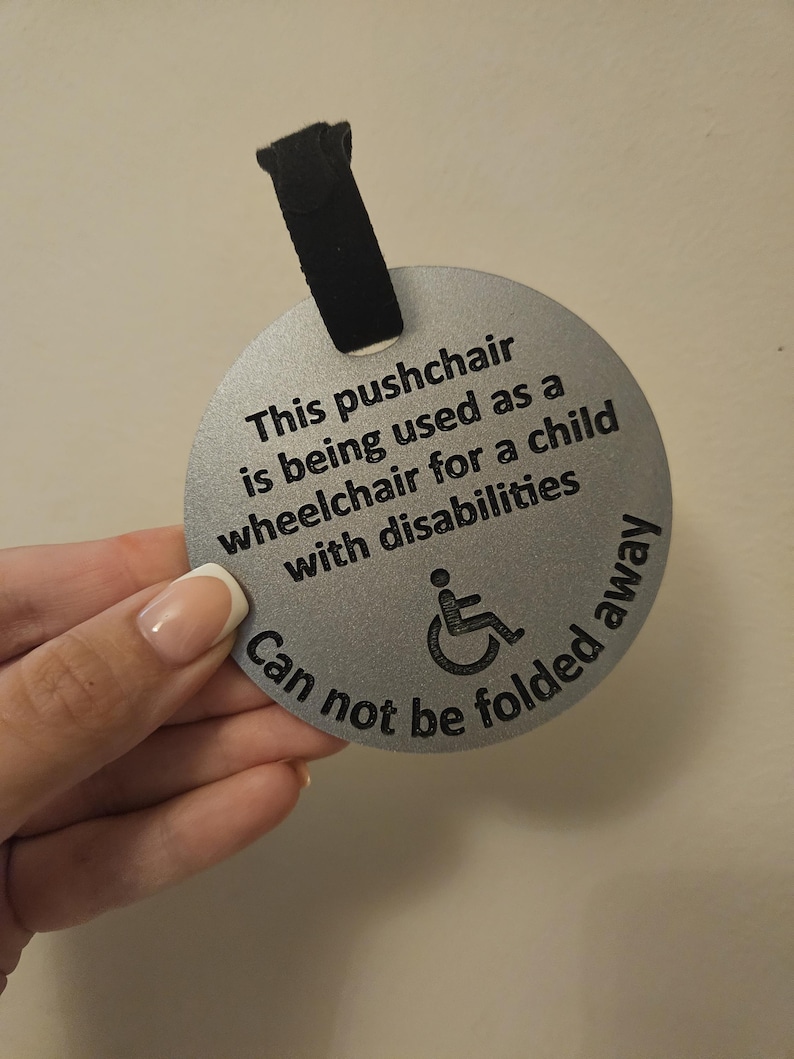 May include: A silver, circular tag with black text and a wheelchair symbol. The text reads: "This pushchair is being used as a wheelchair for a child with disabilities. Can not be folded away."