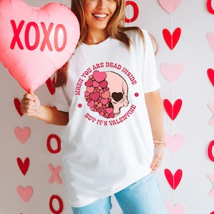 May include: White t-shirt with a skull graphic and the text "When you are dead inside but it's Valentine". The skull is pink and surrounded by pink hearts. A pink heart-shaped balloon with "XOXO" is held in the image.