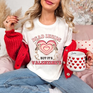 May include: White t-shirt with the text "DEAD INSIDE BUT IT'S VALENTINE'S" in pink and beige. The graphic features a heart with skeletons. A white mug with red hearts and the word "Love" is also visible.
