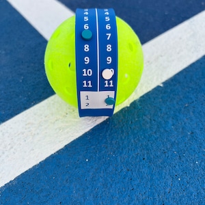 Pode incluir: Uma bola de pickleball amarelo brilhante com um marcador de pontua&ccedil;&atilde;o azul enrolado. O marcador exibe n&uacute;meros de 1 a 11, com marcadores correspondentes. A bola est&aacute; em uma superf&iacute;cie azul com linhas brancas, sugerindo uma quadra de pickleball.