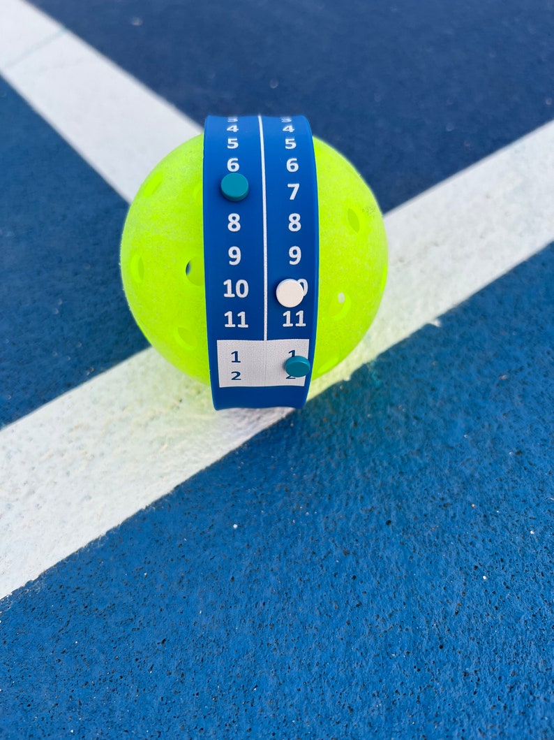 Pode incluir: Uma bola de pickleball amarelo brilhante com um marcador de pontua&ccedil;&atilde;o azul enrolado. O marcador exibe n&uacute;meros de 1 a 11, com marcadores correspondentes. A bola est&aacute; em uma superf&iacute;cie azul com linhas brancas, sugerindo uma quadra de pickleball.
