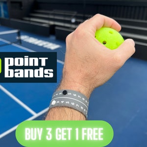 Pode incluir: Um bra&ccedil;o usando uma pulseira Point Bands cinza com n&uacute;meros brancos e um mostrador preto, segurando uma bola de pickleball verde neon. O logotipo Point Bands &eacute; vis&iacute;vel. O texto diz "BUY 3 GET 1 FREE".