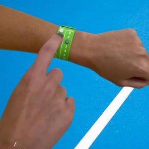 Pode incluir: Uma pulseira verde com n&uacute;meros brancos e fecho de bot&atilde;o &eacute; usada no pulso. Um dedo pressiona o bot&atilde;o. O fundo &eacute; azul com uma faixa branca. A pulseira parece ser um acess&oacute;rio esportivo ou para eventos.