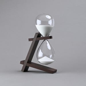 May include: An hourglass with clear glass bulbs containing white sand, supported by a dark brown wooden frame. The frame features an angled base and a horizontal support arm that holds the glass hourglass in a suspended, tilted position.