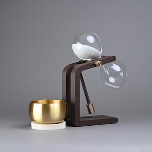May include: A modern coffee brewing device with a dark brown wooden frame, two clear glass globes, and a brass-colored metal bowl. The top globe contains white powder. The bowl sits on a white coaster.