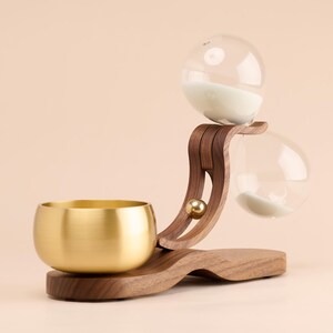 May include: A decorative hourglass with a wooden frame and a brass bowl. The hourglass features two clear glass globes connected by a curved wooden support. The base is also made of wood. The bowl is gold-colored.