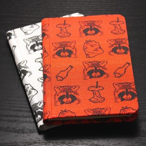 Puede incluir: Dos cuadernos, uno blanco y otro naranja, con patrones de mapaches y basura. El cuaderno blanco tiene caras de mapaches negros. El cuaderno naranja presenta mapaches negros, corazones de manzana y botellas. Los cuadernos están sobre una superficie oscura.