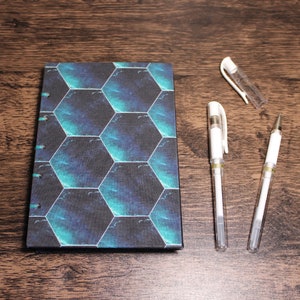May include: A small, blue and black notebook with a hexagonal pattern on the cover. Three white pens are also pictured on a wooden surface.