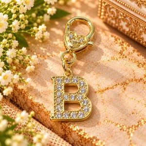 May include: A gold-toned metal charm in the shape of the letter B, encrusted with clear, sparkling rhinestones. The charm features a heart-shaped lobster clasp at the top, also accented with small, clear rhinestones, attached by a circular ring.