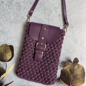 May include: A purple crocheted crossbody bag with a leather flap and buckle closure. The bag has a long strap and is photographed against a gray background.