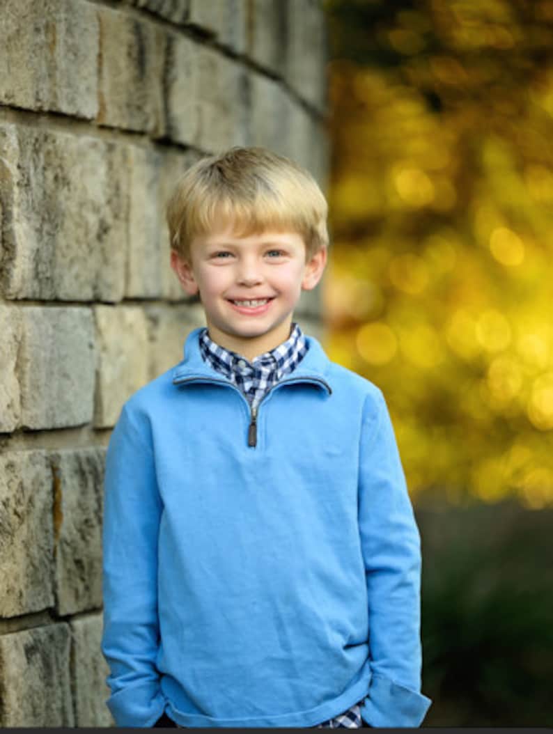 May include: A young boy with blond hair is wearing a light blue quarter-zip sweater over a white and blue checked shirt. He is smiling and standing in front of a stone wall.