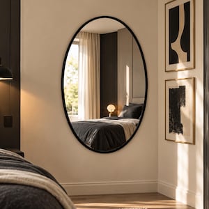 May include: An oval wall mirror with a thin black frame mounted on a light-colored wall. The mirror reflects a bedroom interior featuring a bed with dark bedding, a bedside lamp, and a window with light-colored curtains.