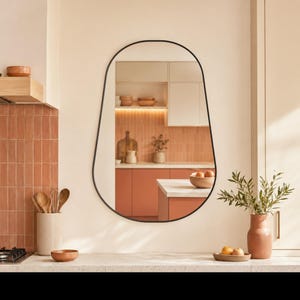 Pode incluir: Um espelho de parede com uma moldura fina, preta e assimétrica, apresentando uma forma orgânica arredondada. A superfície refletora mostra o interior de uma cozinha com azulejos cor de terracota, armários de madeira e uma bancada.