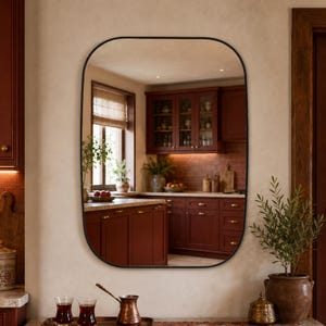 Op de afbeelding: Een rechthoekige wandspiegel met afgeronde hoeken en een dun zwart frame. Het reflecterende oppervlak toont een keukeninterieur met donkerrode kasten, een raam met jaloezieën, een plant in een pot en een aanrecht met diverse keukenitems.