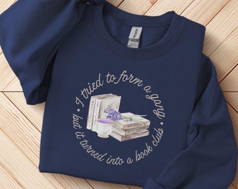 Puede incluir: Una sudadera azul marino con un diseño gráfico blanco de una pila de libros, una taza de té y flores de lavanda. El texto en el gráfico dice "Traté de formar una pandilla, pero se convirtió en un club de lectura."