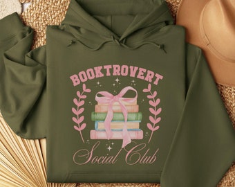 May include: A green hooded sweatshirt with the text "Booktrovert Social Club" in pink lettering. The design features a stack of books tied with a pink ribbon and surrounded by pink leaves.