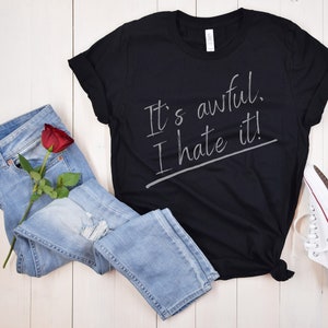 Puede incluir: Camiseta negra con el texto "It's awful. I hate it!" escrito en blanco en la parte delantera. La camiseta está colocada sobre una superficie de madera blanca con un par de pantalones vaqueros azules y una rosa roja.