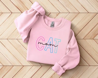 以下が含まれることがあります： レインボーカラーの「CAT mom」というグラフィックデザインが施されたピンク色のスウェットシャツです。