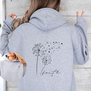Pu&ograve; includere: Felpa con cappuccio grigia con un design a dente di leone e la parola "faith" stampata sul retro.