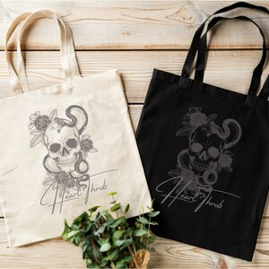 Pode incluir: Duas sacolas de tecido com um design de caveira e cobra. A sacola à esquerda é branca com texto preto e a sacola à direita é preta com texto branco. Ambas as sacolas têm o texto "Heart Throb".