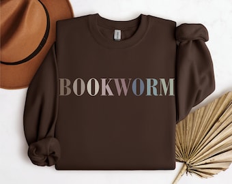 Puede incluir: Una sudadera marrón oscuro con la palabra "BOOKWORM" impresa en el pecho en un degradado de colores pastel. La sudadera se muestra con un sombrero marrón y una hoja de palma seca.
