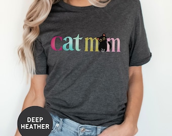 Può includere: Una maglietta grigio antracite con il testo "cat mom" in colori arcobaleno. Un gatto nero con gli occhi bianchi è seduto nella "m" di "mom".