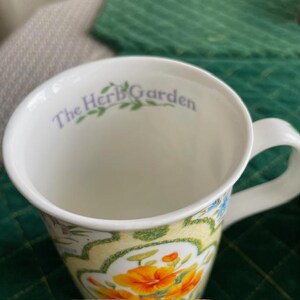 May include: White ceramic mug with a floral design featuring orange and yellow flowers, green leaves, and blue accents. The words "The Herb Garden" are printed in light purple inside the rim of the mug.