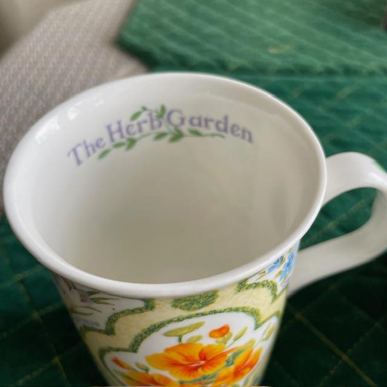 May include: White ceramic mug with a floral design featuring orange and yellow flowers, green leaves, and blue accents. The words "The Herb Garden" are printed in light purple inside the rim of the mug.