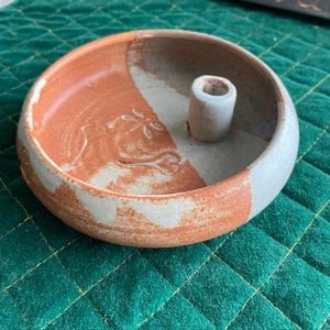 May include: A handmade ceramic candle holder with a circular design. The holder features a light beige and reddish-brown color scheme, with a raised floral design on the reddish-brown section. A small, cylindrical holder is in the center.