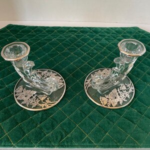 May include: Two clear glass candle holders with silver rims and floral designs. The candle holders have a curved shape and sit on round bases with silver floral patterns. The bases are approximately 4 inches in diameter.