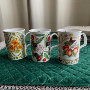 May include: Three white ceramic mugs with decorative floral and fruit designs. One mug features orange flowers, another has cherries and leaves, and the third displays peaches and a butterfly. Each mug has a white handle.